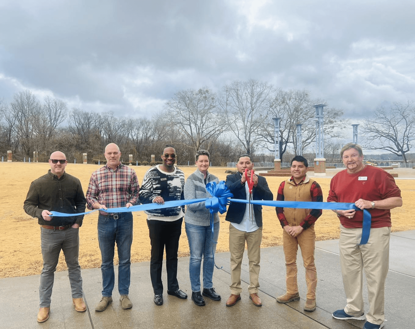 A group gathered outdoors for a ribbon cutting, holding a blue ribbon while one participant holds ceremonial scissors